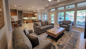 A spacious living room with a sofa set, a coffee table, and a TV. There is a kitchen area in the background with wooden cabinets and a dining table.