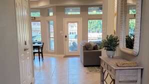 A spacious hallway with a tiled floor, a grey sofa, a console table with a mirror, and a door leading to a dining area.