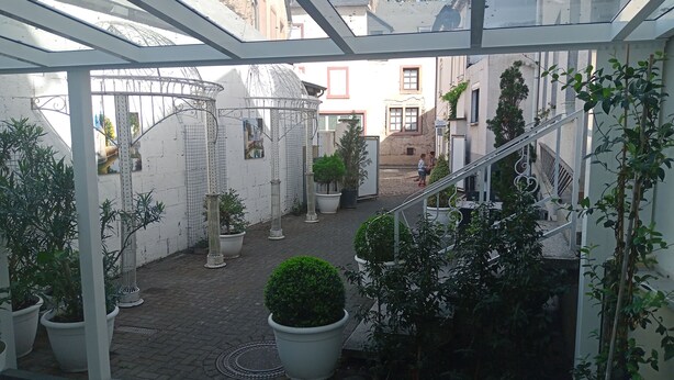 A covered courtyard with potted plants, a metal staircase, and a view of buildings in the background. - New apartment at the market place Lieser Moseloefer 30 meters (Lieser)