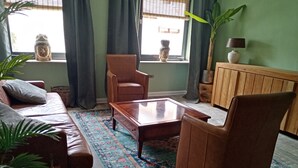 A living room with a brown leather sofa, a wooden coffee table, and a patterned rug. There are two windows with curtains, a potted plant, and a lamp on a wooden cabinet. - New apartment at the market place Lieser Moseloefer 30 meters (Lieser)