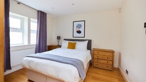 A bedroom with a bed, a nightstand, a dresser, a window with curtains, and a wall-mounted artwork. - The Bayswater Nook - Stylish 1bdr Flat (London)