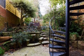 Un chemin de pierre menant à un escalier en colimaçon, entouré de verdure et bordé d’un petit étang.