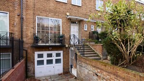 A brick building with a white garage door, a small balcony with a metal railing, and a staircase leading to the entrance. - The Bayswater Nook - Stylish 1bdr Flat (London)