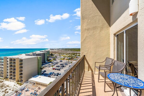 Un balcon avec une table et des chaises, donnant sur une plage et un parking.