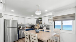 Een moderne keuken met witte kasten, een roestvrijstalen koelkast, een houten eettafel met vier stoelen en uitzicht op de oceaan door grote ramen.