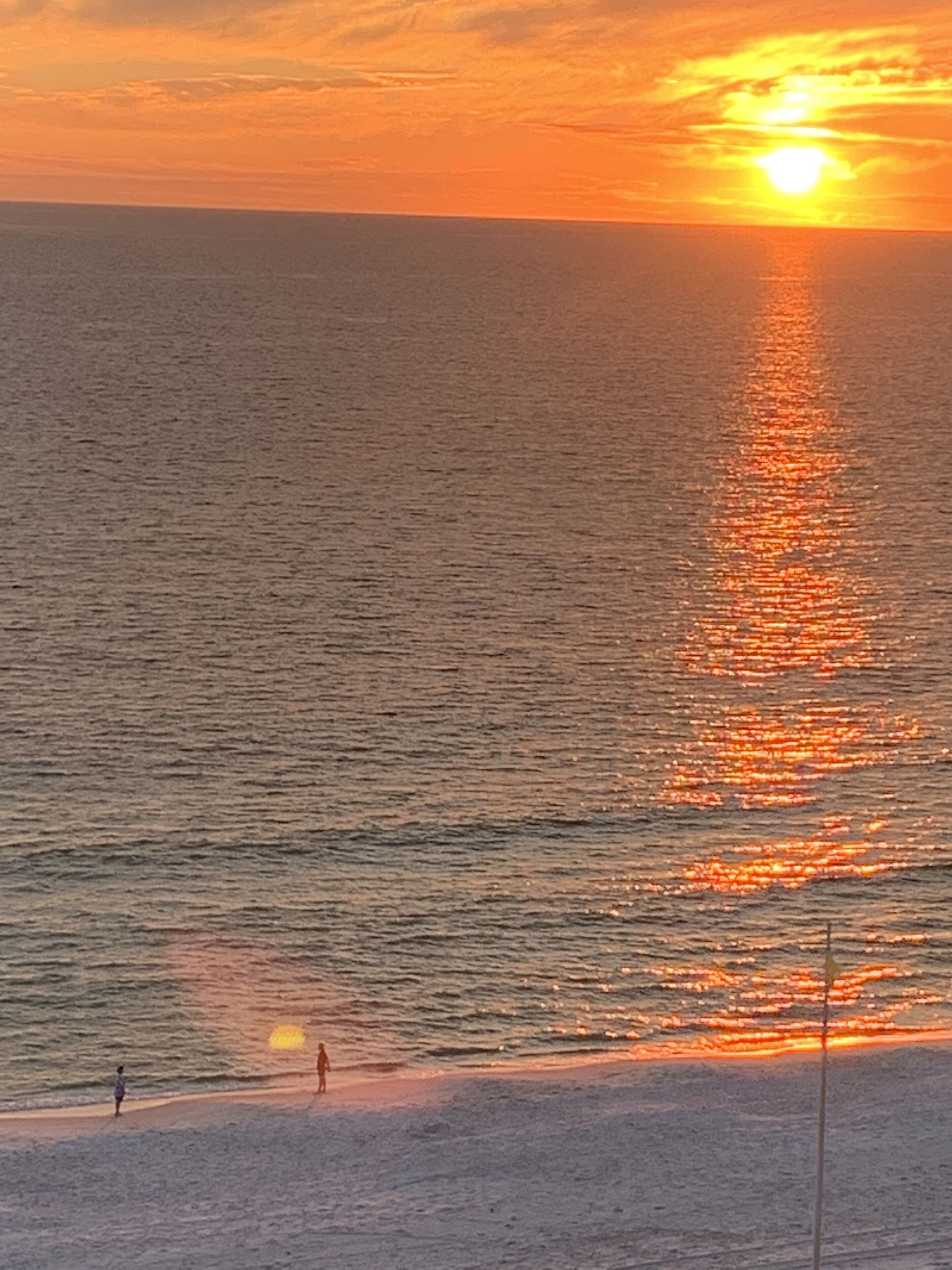 A beach scene with the sun setting over the ocean, creating a reflection on the water.
