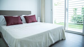 Ein ordentlich bezogenes Bett mit weißer Bettwäsche und zwei roten Kissen, ein grauer Kopfteil und Blick auf Grün durch eine Schiebeglastür.
