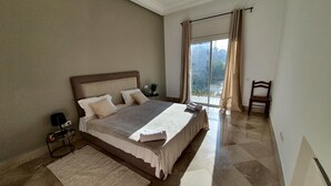 Ein geräumiges Schlafzimmer mit einem großen Bett, einem kleinen Nachttisch, einem Sessel und Blick ins Freie durch ein großes Fenster.