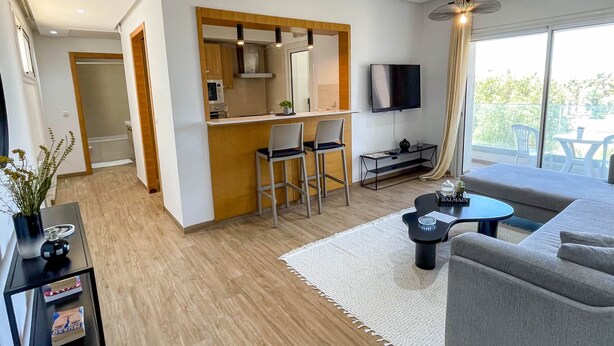 Una sala de estar moderna con un sofá gris, una mesita negra pequeña y un tapete blanco. Cuenta con una zona de cocina con armarios de madera y un televisor montado en la pared.