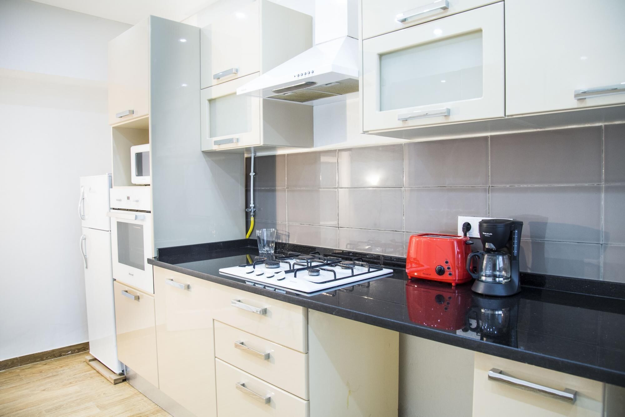 Une cuisine moderne avec des meubles blancs, un plan de travail noir, une cuisinière à gaz, un grille-pain rouge et une machine à café.