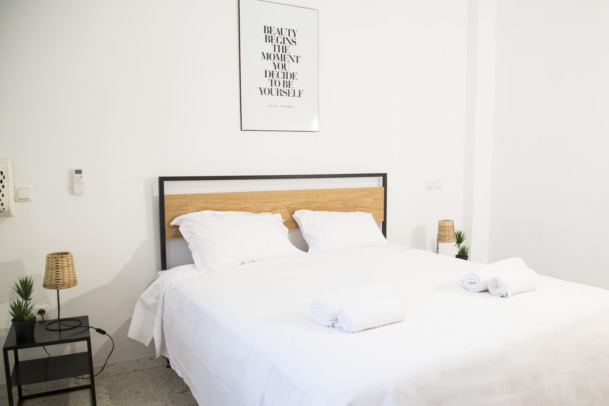 A neatly made bed with white linens, a wooden headboard, and two white pillows. There is a small bedside table with a lamp and a potted plant. A framed quote is hanging on the wall above the bed.