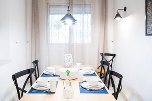 Un coin repas avec une table en bois dressée pour quatre personnes, des chaises noires, un luminaire suspendu et une fenêtre ornée de barreaux en fer décoratifs.