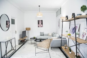 Un salon moderne avec un canapé gris, un fauteuil en osier, une table basse et un téléviseur. On peut y voir des tableaux encadrés au mur, une bibliothèque et une plante en pot.
