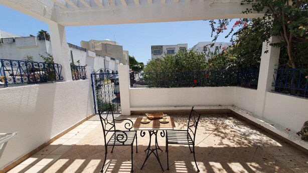 En overdækket terrasse med et bord og stole, en pergola over hovedet, og udsigt til bygninger og grøn vegetation i baggrunden.