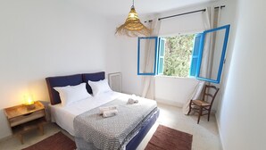 Un dormitorio ordenado con cama, mesita de noche, lámpara y ventana con contraventanas azules.