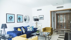 Ein modernes Wohnzimmer mit einem blauen Sofa, gelben Sessel und einem Couchtisch. An der Wand hängen gerahmte Bilder und auf einem Beistelltisch steht eine Lampe.