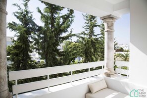 Un balcón con un banco blanco, una columna de piedra y árboles al fondo.
