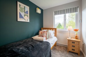 A bedroom with a bed, a bedside table with a lamp and a vase, a radiator, and a window with curtains.