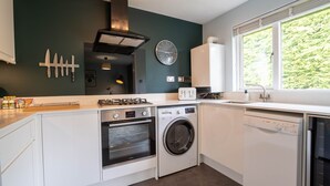 Una cocina moderna con horno empotrado, lavadora y lavavajillas, un reloj en la pared y una ventana con vista a zonas verdes.