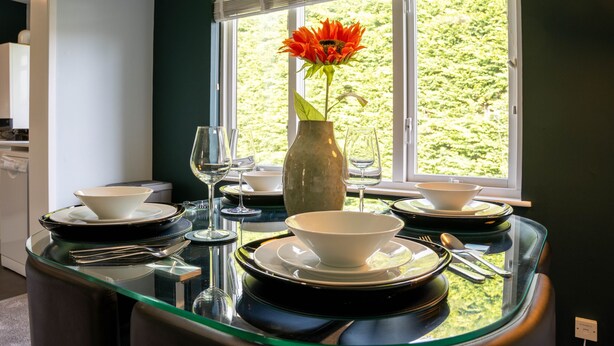 Una mesa de comedor dispuesta con cuencos, platos y cubiertos blancos, un florero con una sola flor, y una copa de vino.