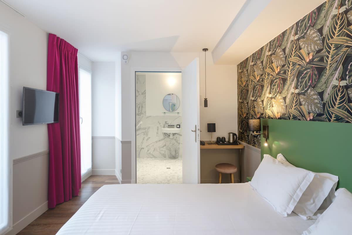 A hotel room with a bed, a desk, a television, and a bathroom visible through an open door.