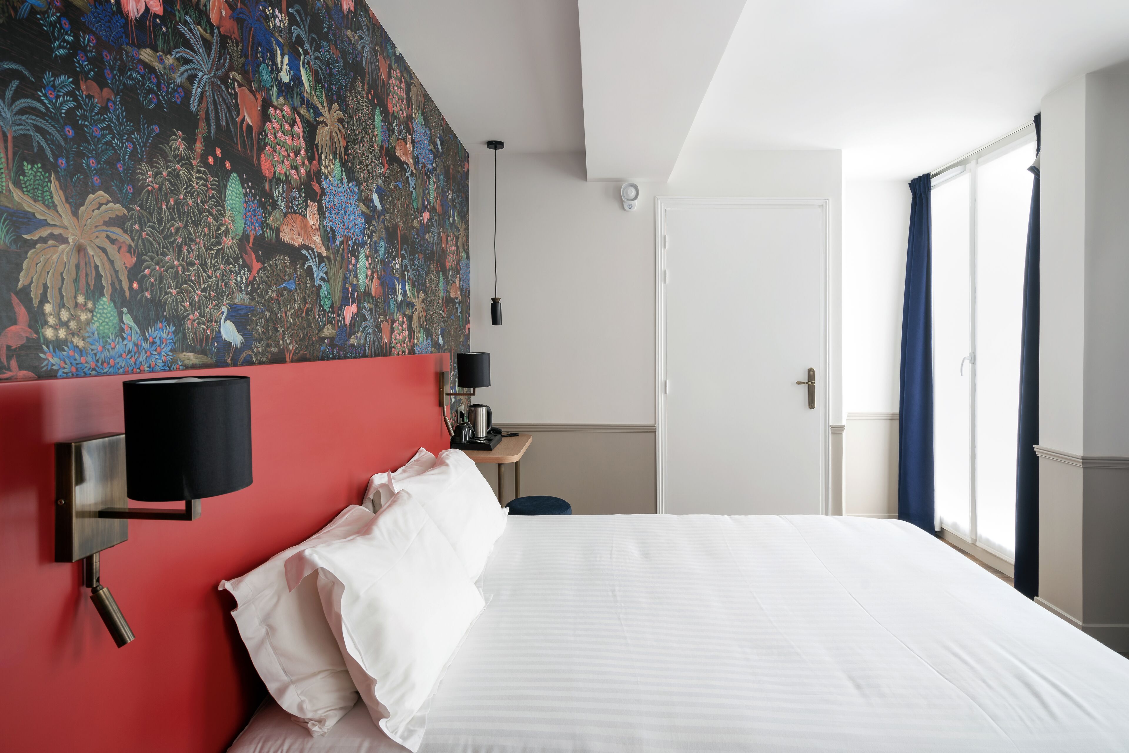 A hotel room with a large bed, a red accent wall, a wall-mounted lamp, a desk with a chair, and a window with curtains.