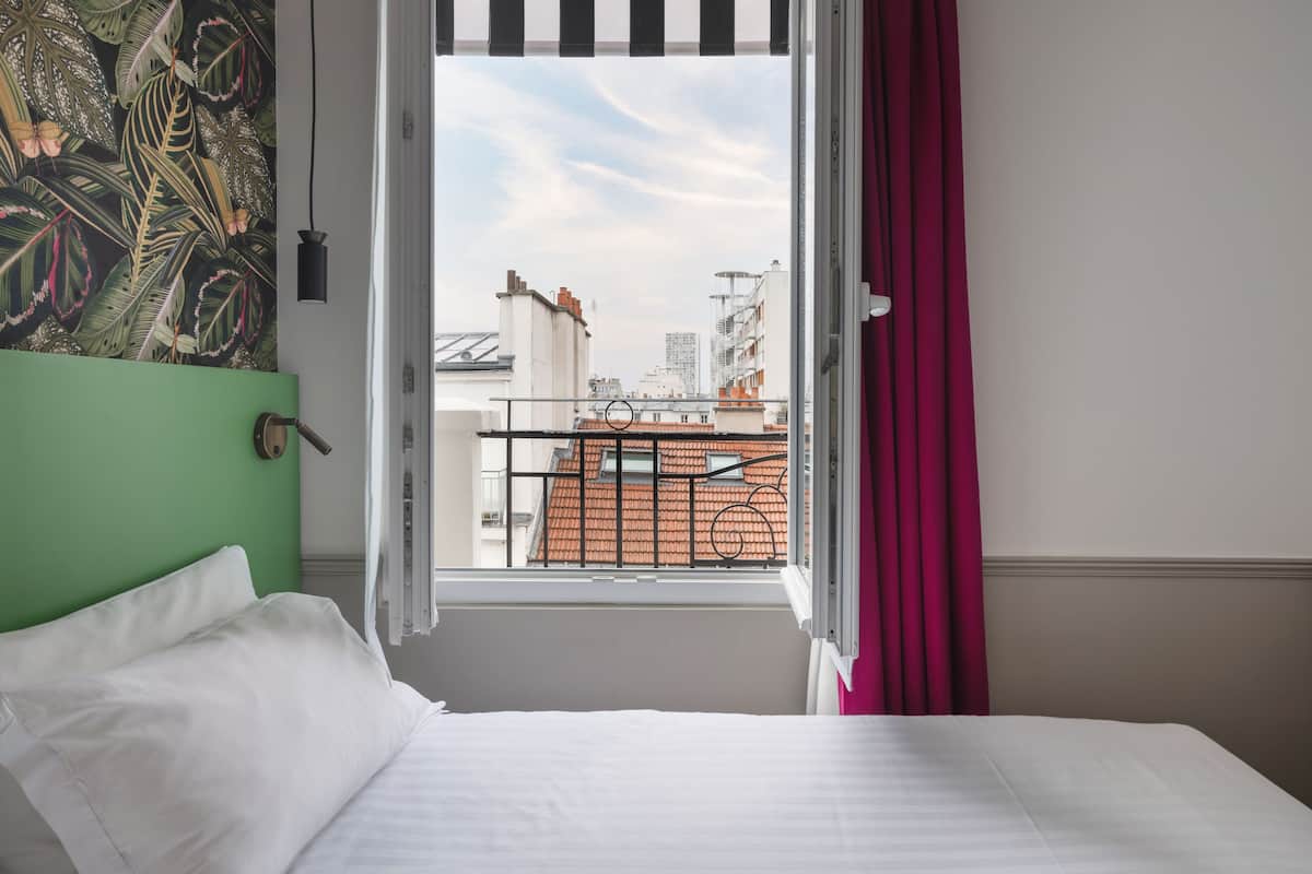 A hotel room with a bed, a window with curtains, a view of buildings, and a green wall.