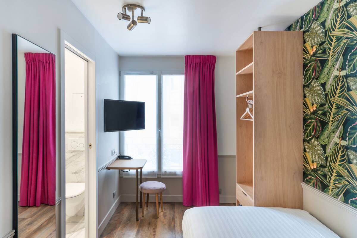 A bedroom with a bed, a TV, a wooden wardrobe, a desk with a chair, and a window with curtains.