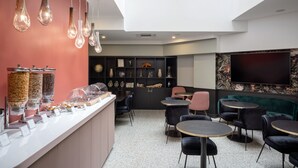 Un área de descanso moderna con techo alto, tragaluces, una barra con diferentes dispensadores de comida y un espacio de asientos con mesas y sillas redondas.