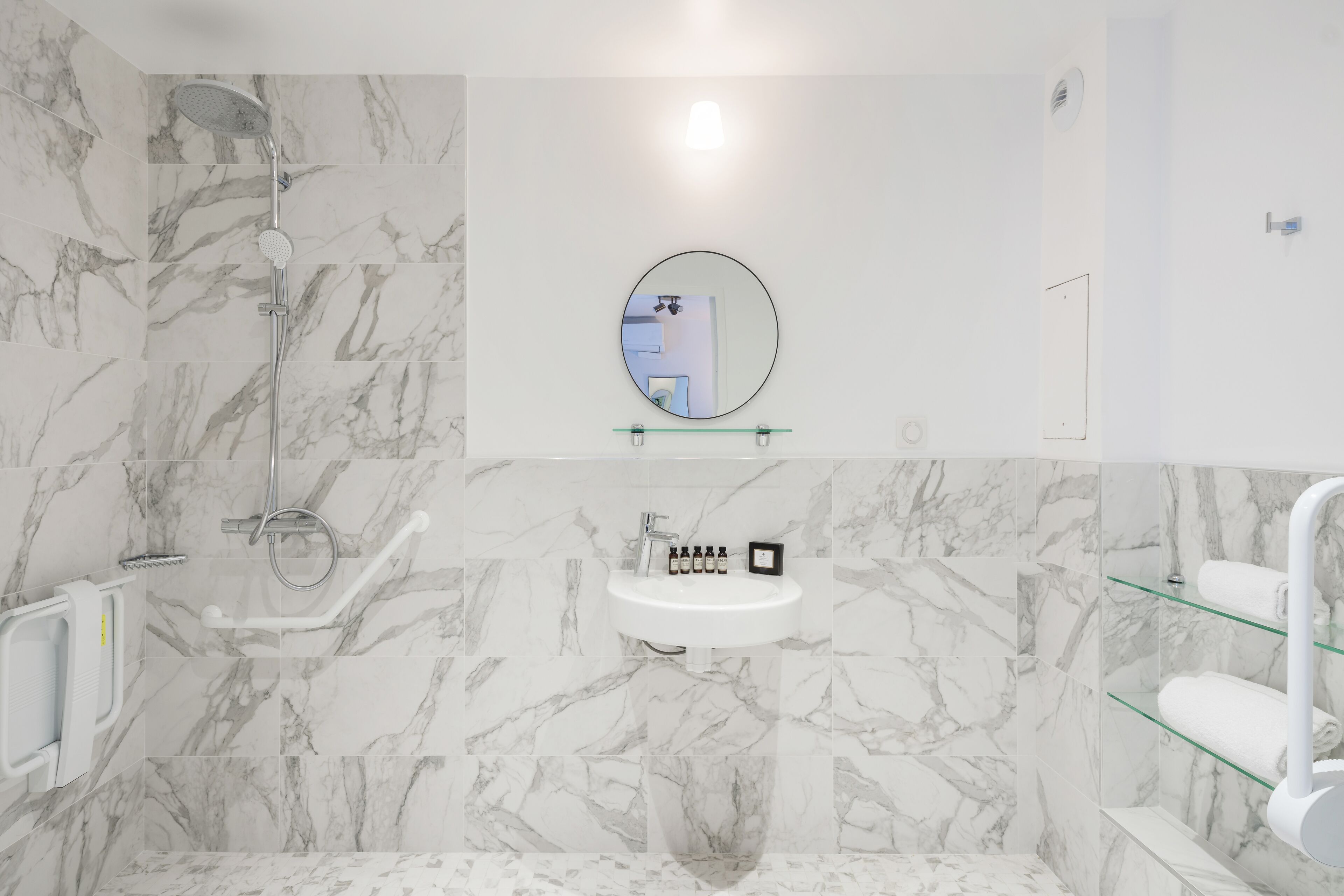 A modern bathroom with a marble wall, a round mirror, a sink, and a glass shelf.