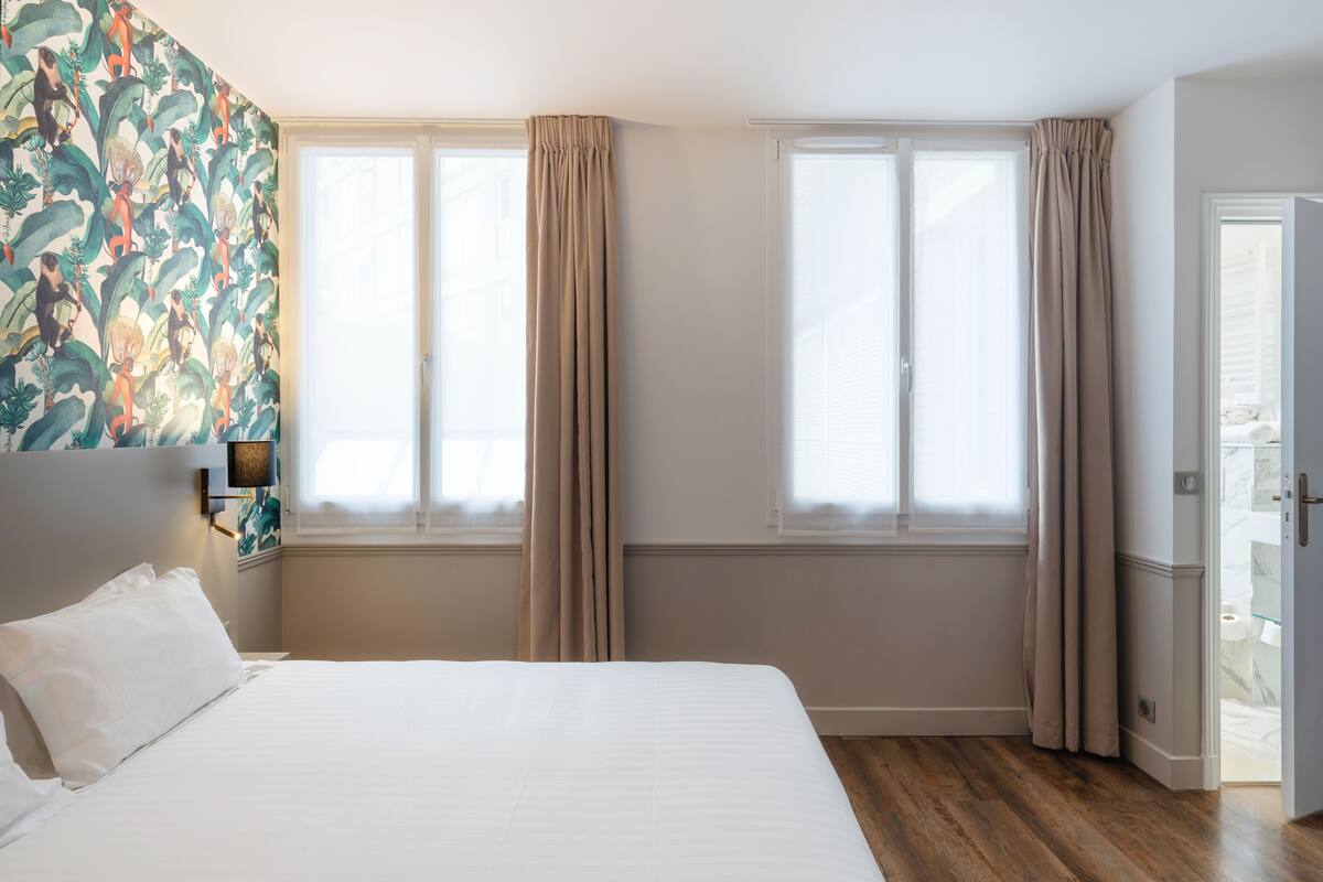 A hotel room with a large bed, two windows with curtains, a headboard with a light fixture, and a door leading to a bathroom.