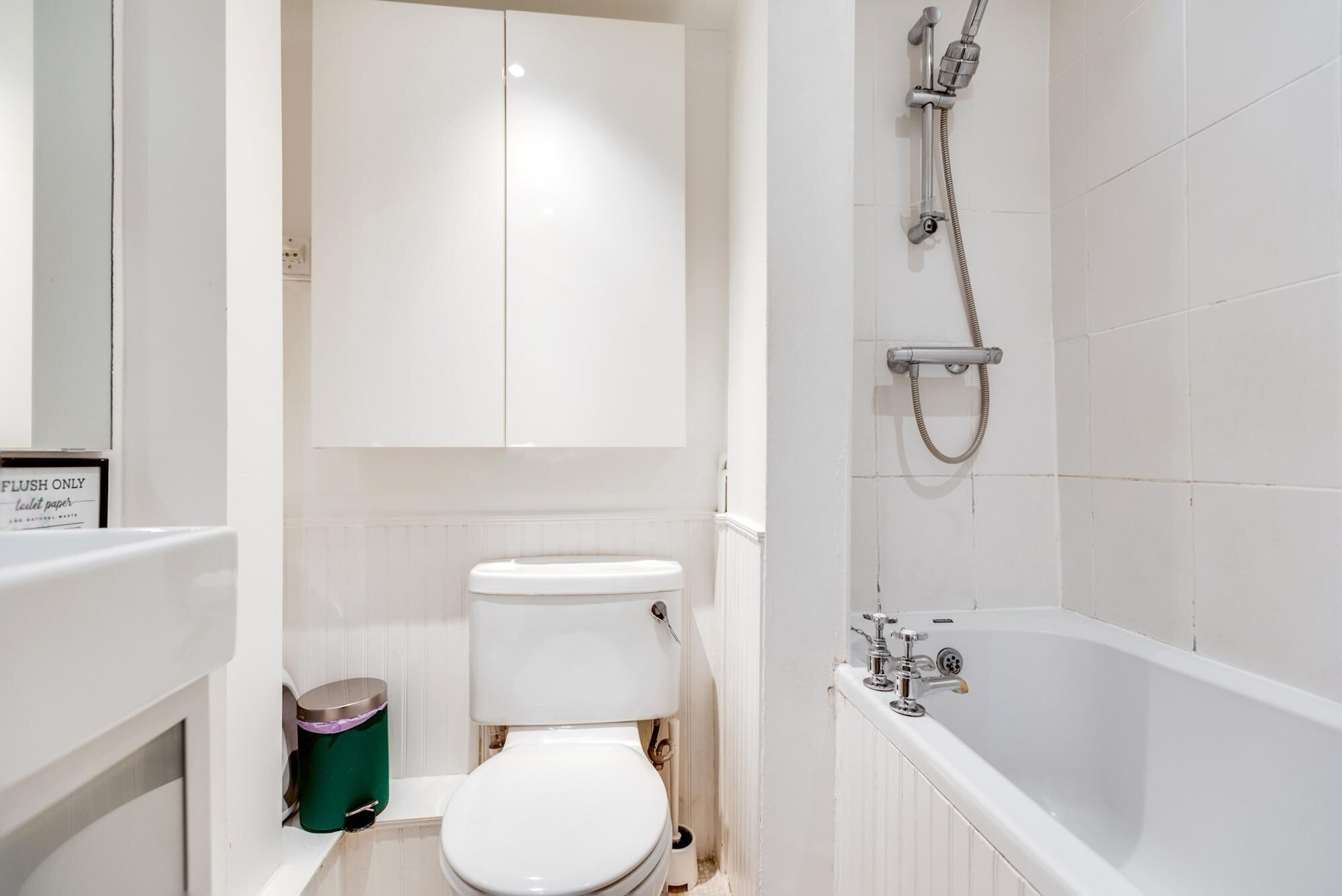 A bathroom with a toilet, a green trash bin, a shower, and a sign that says "FLUSH ONLY toilet paper".