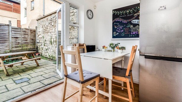 A compact kitchen with a white table, two wooden chairs, a stainless steel refrigerator, and a chalkboard wall with writing. - The St Clements Townhouse 3 Bedroom Bijou Central (Oxford)