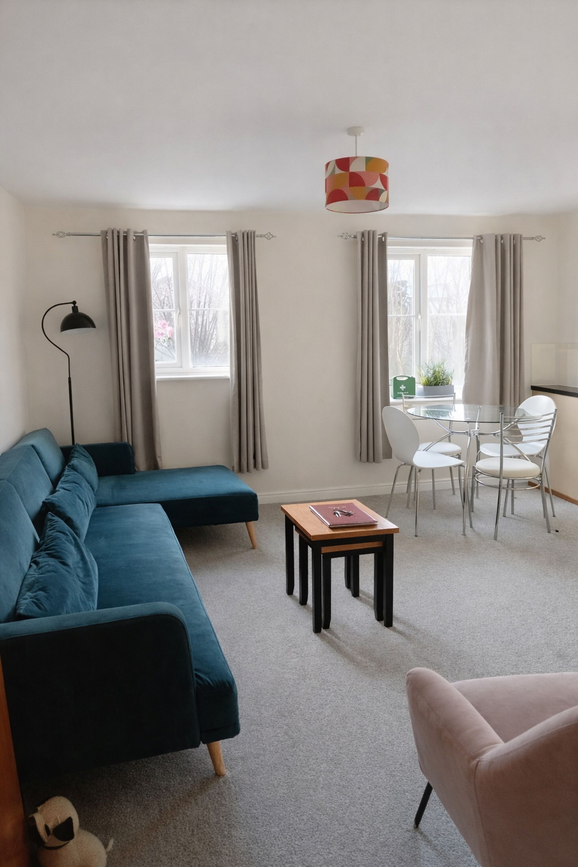 Uma sala de estar moderna com um sofá seccional azul, uma mesinha com um livro, uma cadeira branca e uma mesa de jantar com cadeiras.