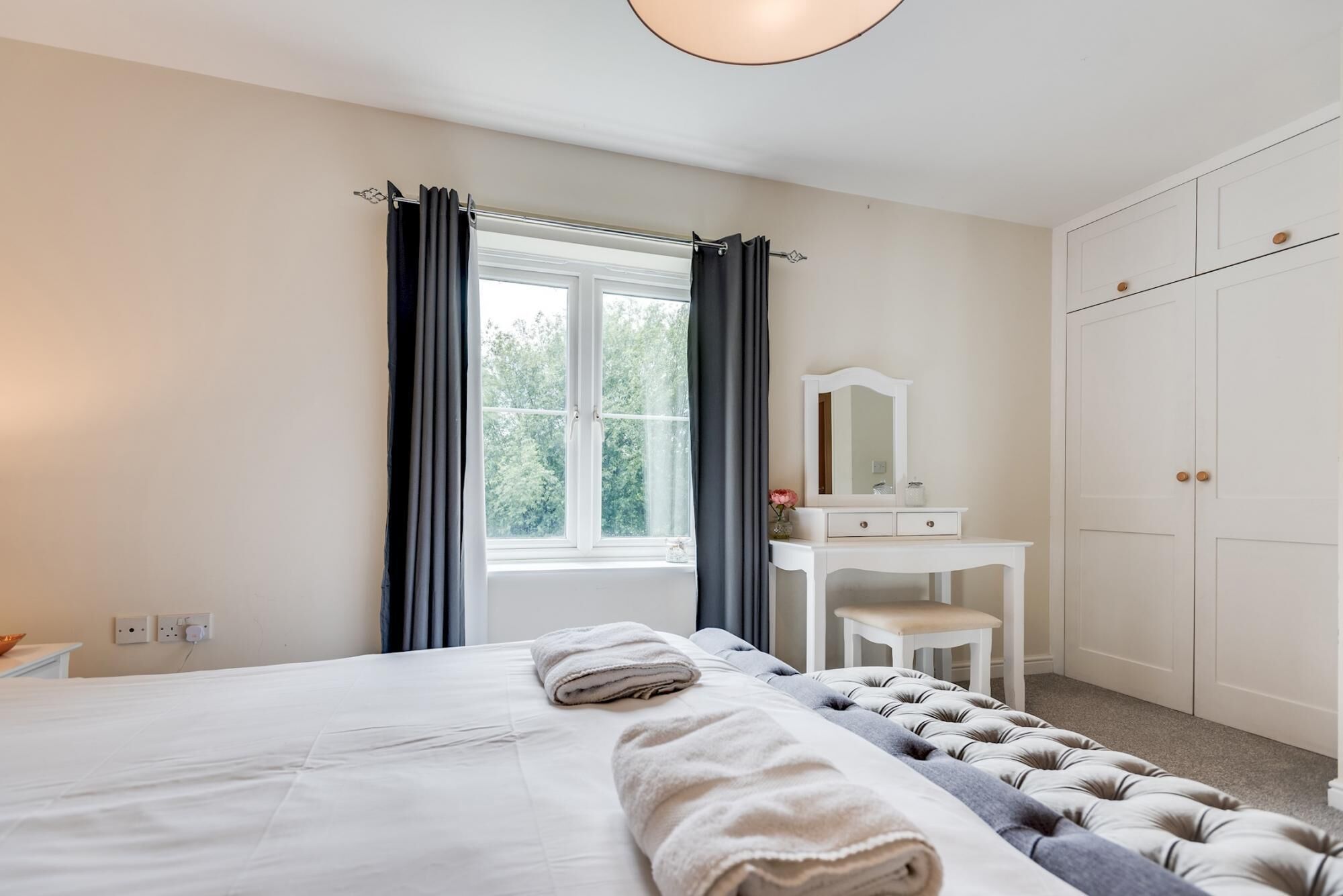 A bedroom with a bed, a vanity with a mirror, a window with curtains, and a wardrobe.