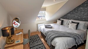 A bedroom with a sloped ceiling, a bed with a grey and white striped blanket, a bedside table with a lamp, and a window with a view of a building. - A Very British Place to Stay - 3 Bedrooms Central (Oxford)