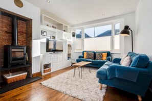 A modern living room with a fireplace, a blue sofa, a TV, and a large window. - Charming 3 BR House - Garden Parking Near CS Lewis Park (Oxford)