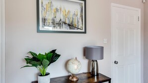 Una pintura enmarco con un paisaje urbano, una planta en maceta, un globo terráqueo y una lámpara sobre una consola de madera, todo ello frente a una pared blanca con una puerta.