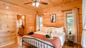 Un dormitorio de cabaña de madera con una cama, una mesita de noche, un espejo y una ventana con cortinas.