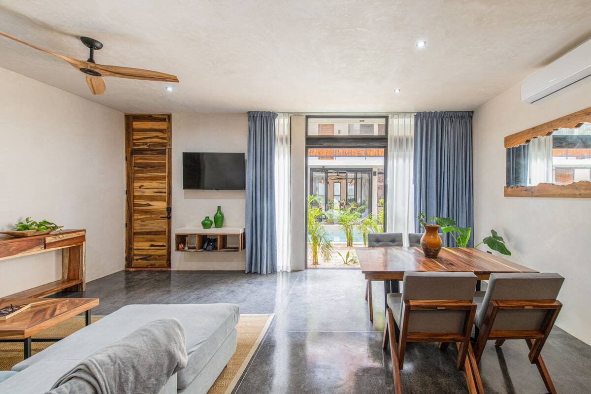 Ein modernes Wohnzimmer mit einer Couch, einem Holzessttisch und Stühlen. Ein Fernseher ist an der Wand montiert, ein Deckenventilator ist vorhanden und es gibt ein großes Fenster mit Vorhängen.