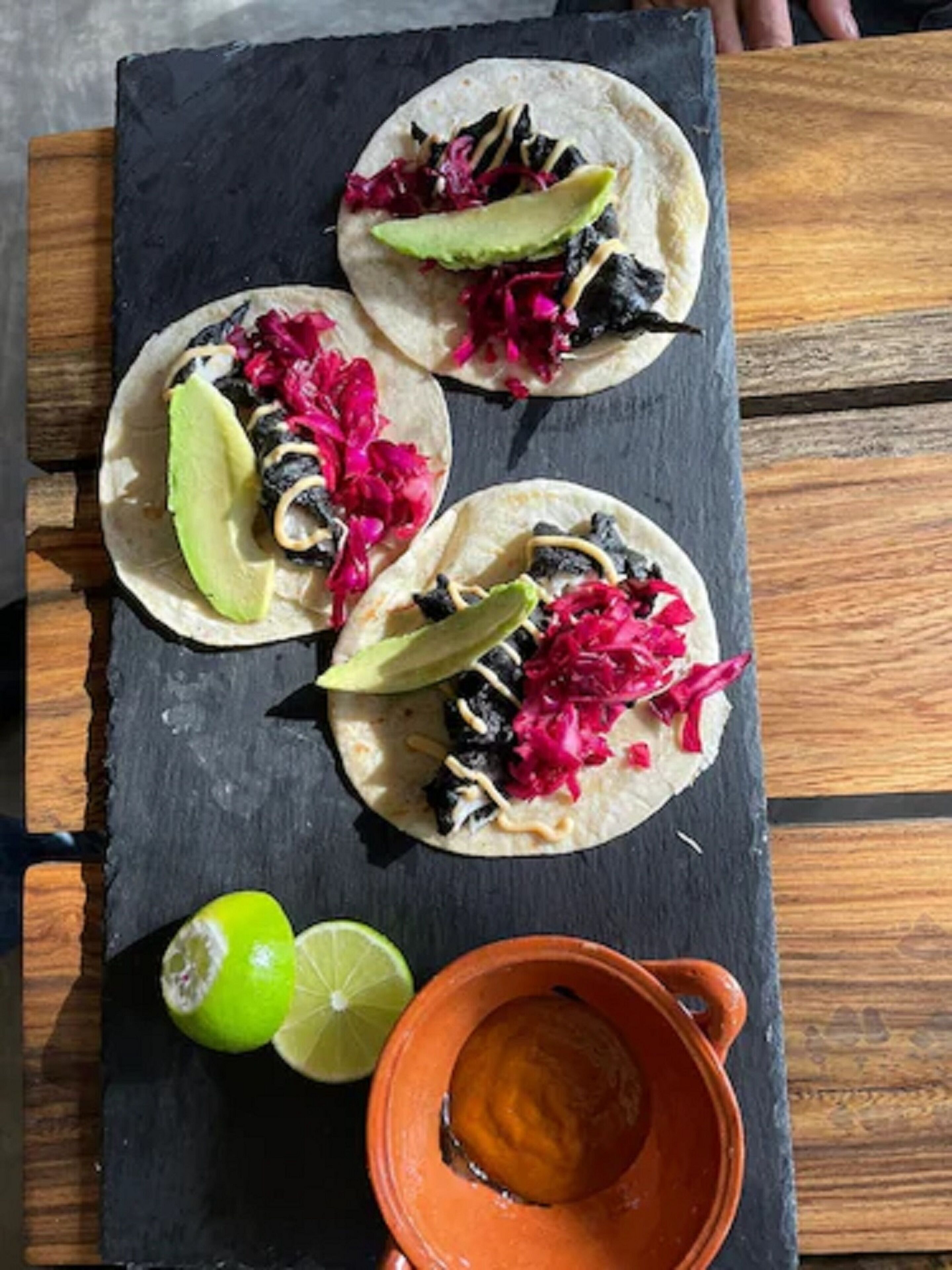 Three tacos on a slate board with lime wedges and a small clay pot.