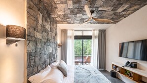 A modern bedroom with a stone wall, a large bed, a ceiling fan, a TV, and a balcony with a view of greenery. - Irie Tulum  (Tulum)