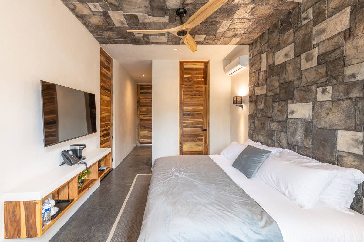 Ein modernes Hotelzimmer mit einer Wand aus Stein, einem großen Bett mit weißer Bettwäsche, einem Flachbildfernseher auf einer Holzkonsole und einem integrierten Schreibtisch mit Telefon.