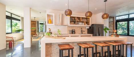 A modern kitchen with a central island, bar stools, pendant lights, and a view of the outdoors.