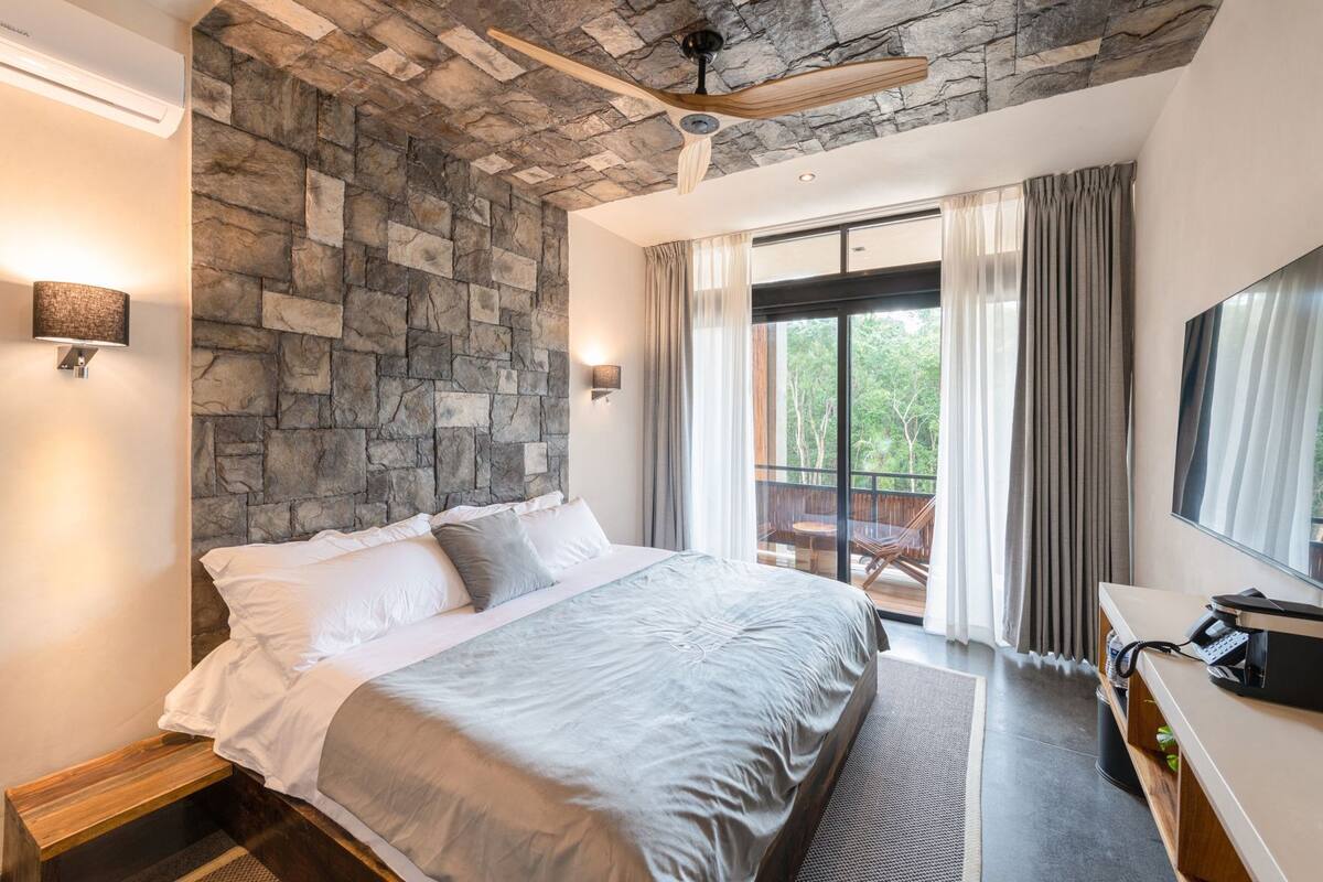Ein Schlafzimmer mit Steingemäuer, einem Bett, einem Deckenventilator, einem Fenster mit Vorhängen und einem Fernseher.