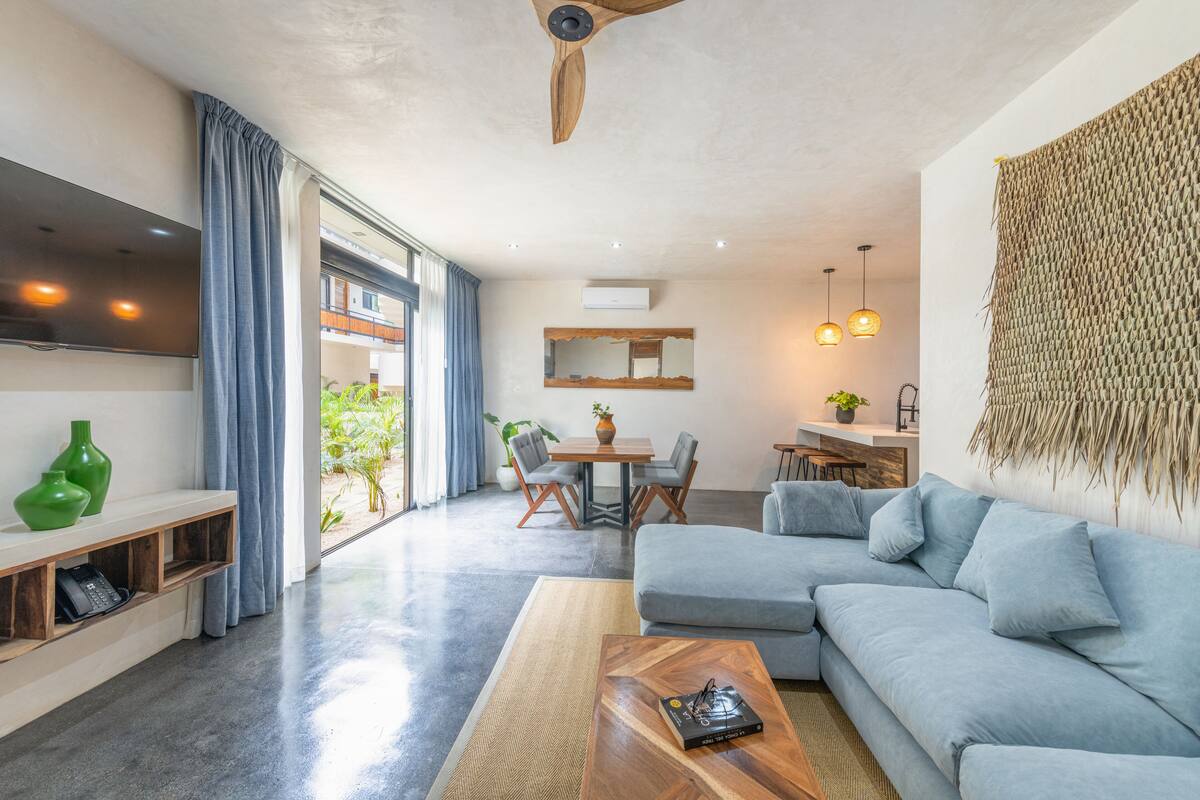 Ein modernes Wohnzimmer mit grauem Sofa, Couchtisch aus Holz und Wand-TV. Ein Essbereich mit Tisch und Stühlen sowie eine Küchenzeile mit Pendelleuchte.