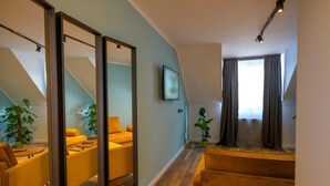 Una sala de estar moderna con un sofá, una mesa de centro y un televisor montado en la pared. Hay dos plantas en macetas y una ventana con cortinas.
