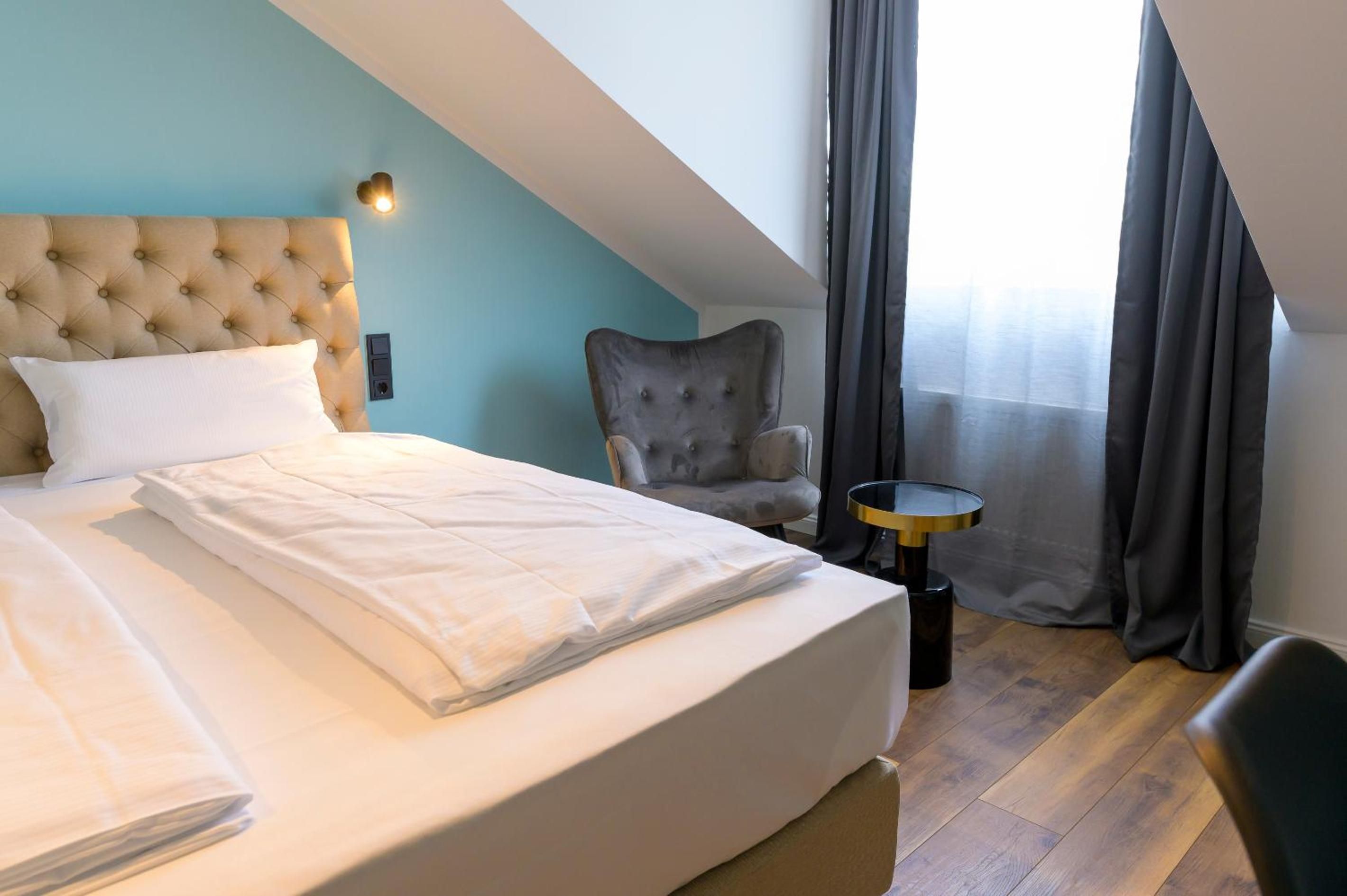 A hotel room with a bed, a chair, a small table, and a window with curtains.