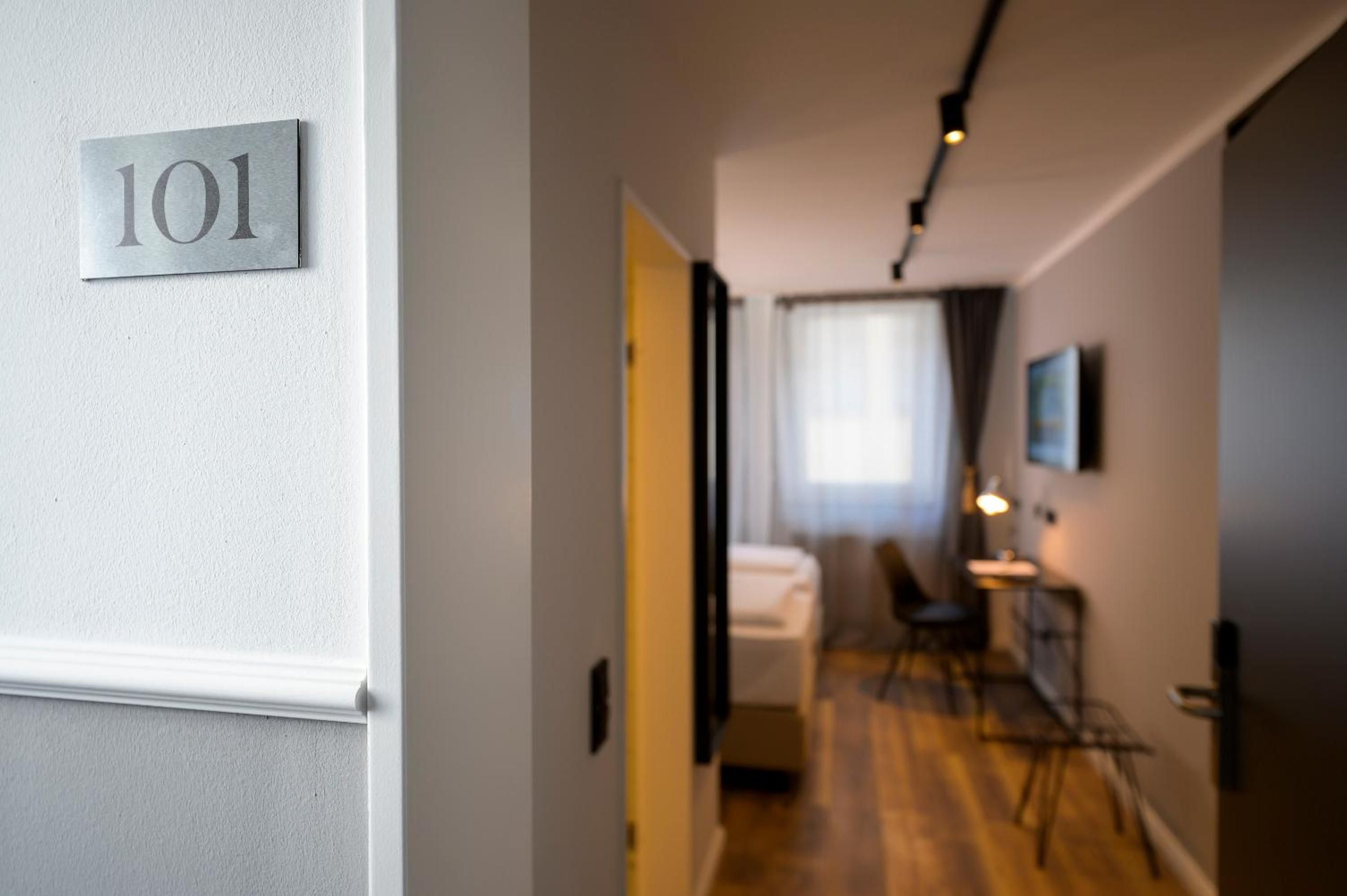 A hotel corridor with a number sign indicating room 101, a bed, a desk, and a chair.