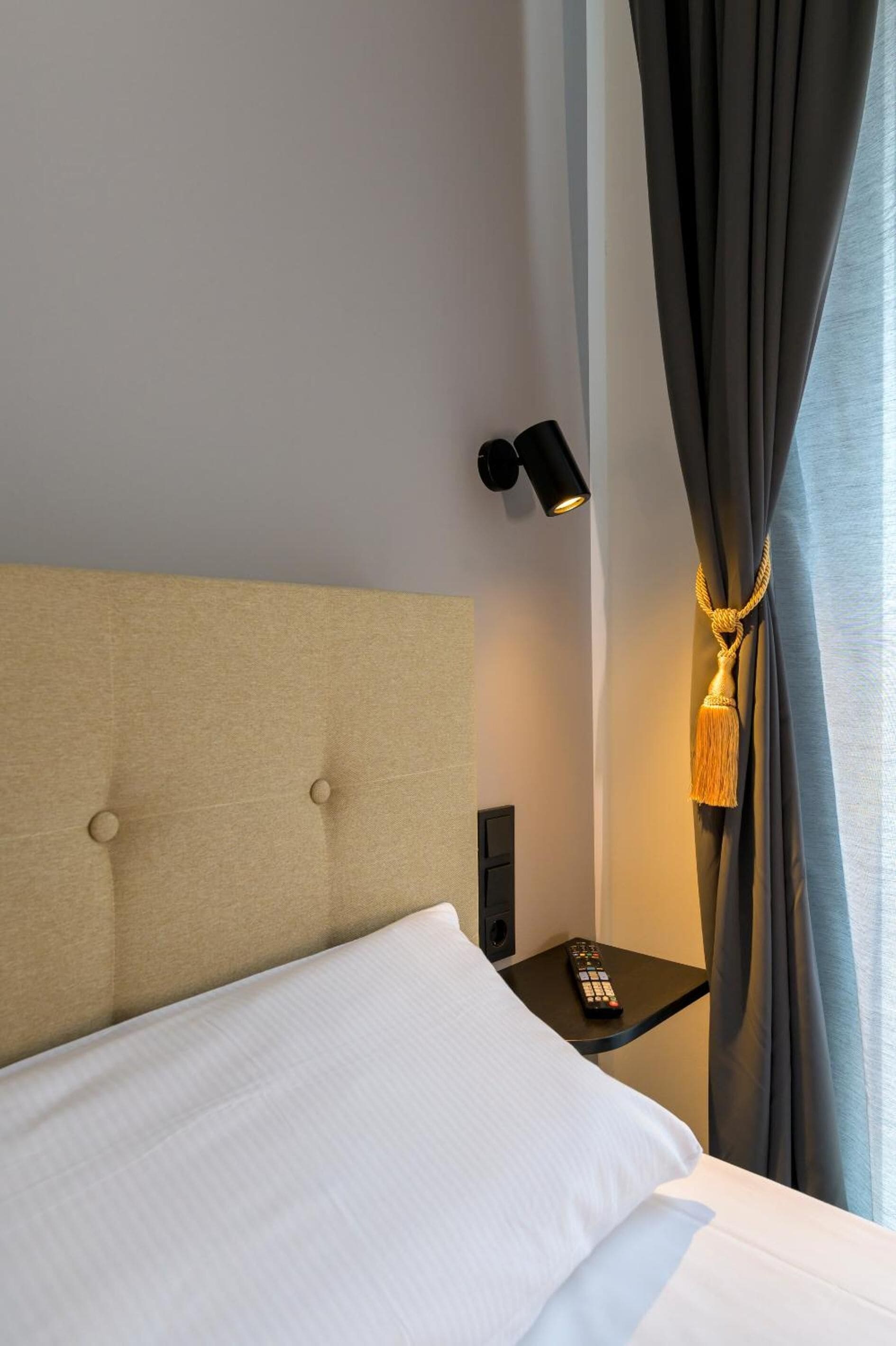 A hotel room with a tufted headboard, a bedside table with a remote control, and a curtain with a tassel.